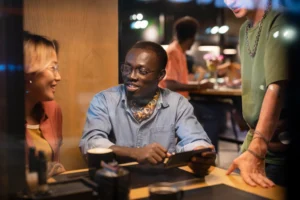 In the image, a diverse group of friends is hanging out together at a dimly lit cafe or restaurant table. A man wearing glasses smiles while holding a smartphone horizontally, showing something to a smiling woman on his left and another friend standing on his right.