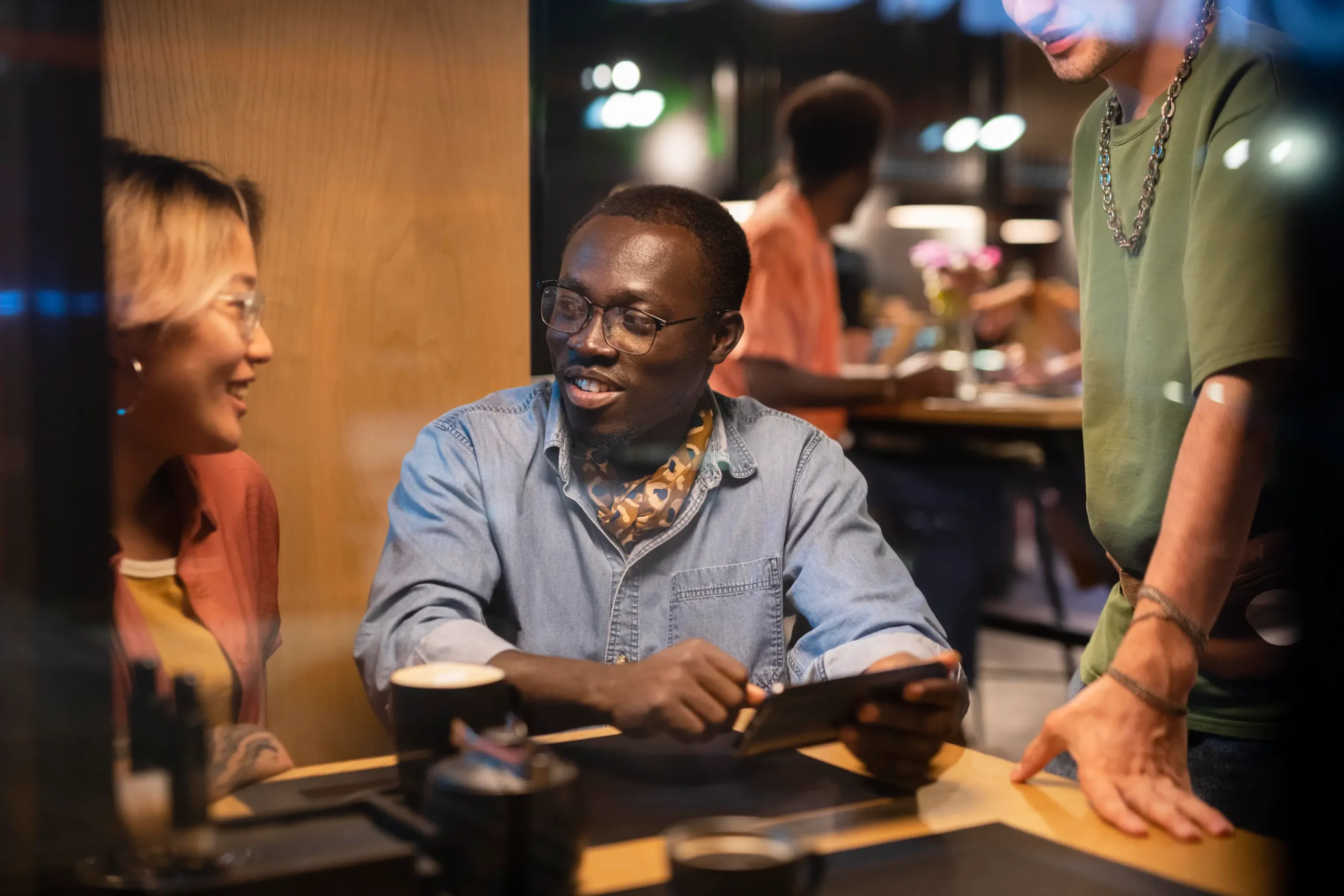 In the image, a diverse group of friends is hanging out together at a dimly lit cafe or restaurant table. A man wearing glasses smiles while holding a smartphone horizontally, showing something to a smiling woman on his left and another friend standing on his right.