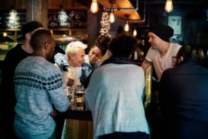 A diverse group of friends gathers around a dimly lit bar counter, smiling and enjoying drinks together under warm hanging pendant lights. They are engaged in cheerful conversation with beer mugs resting on the wooden counter in a cozy pub setting.