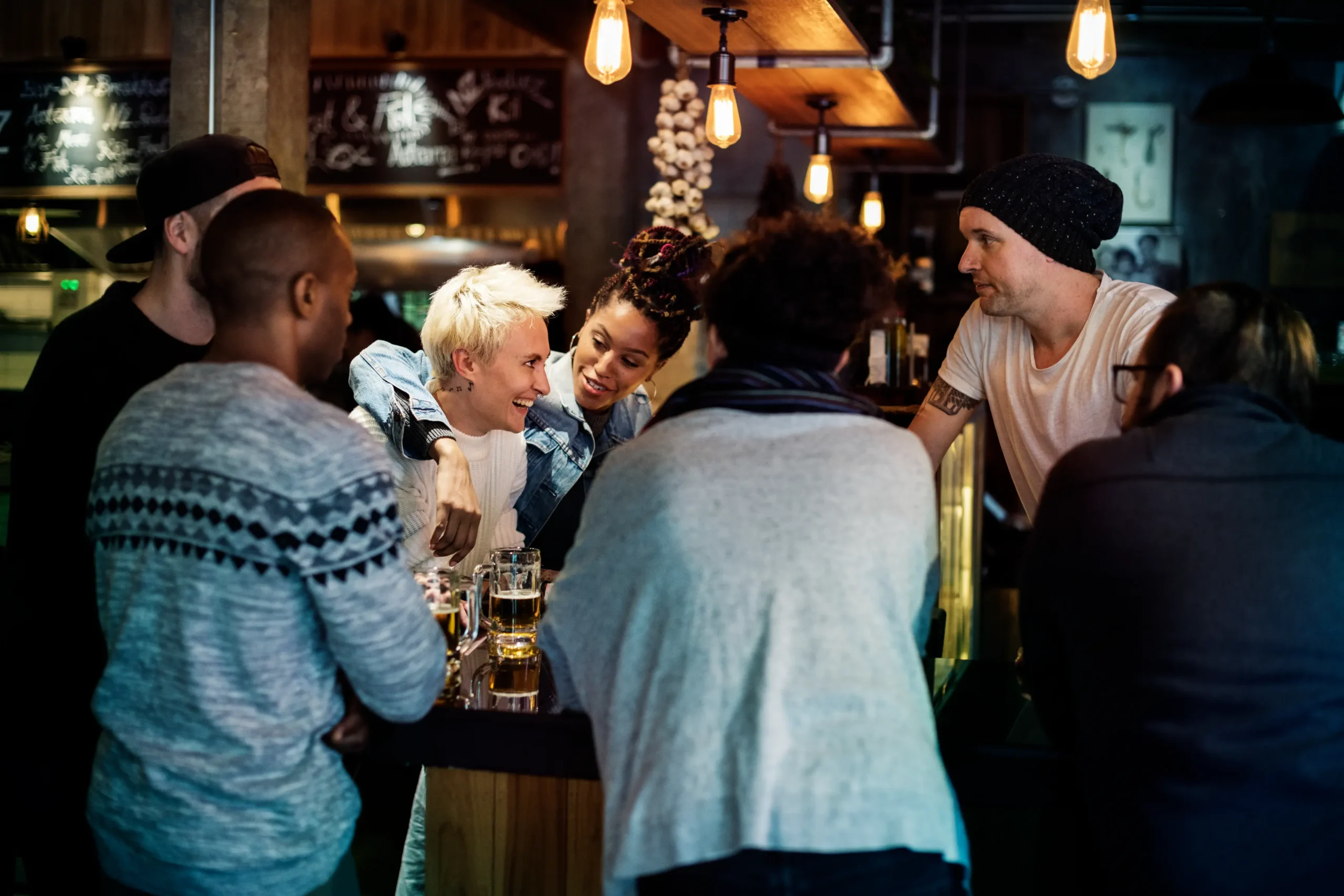 A diverse group of friends gathers around a dimly lit bar counter, smiling and enjoying drinks together under warm hanging pendant lights. They are engaged in cheerful conversation with beer mugs resting on the wooden counter in a cozy pub setting.