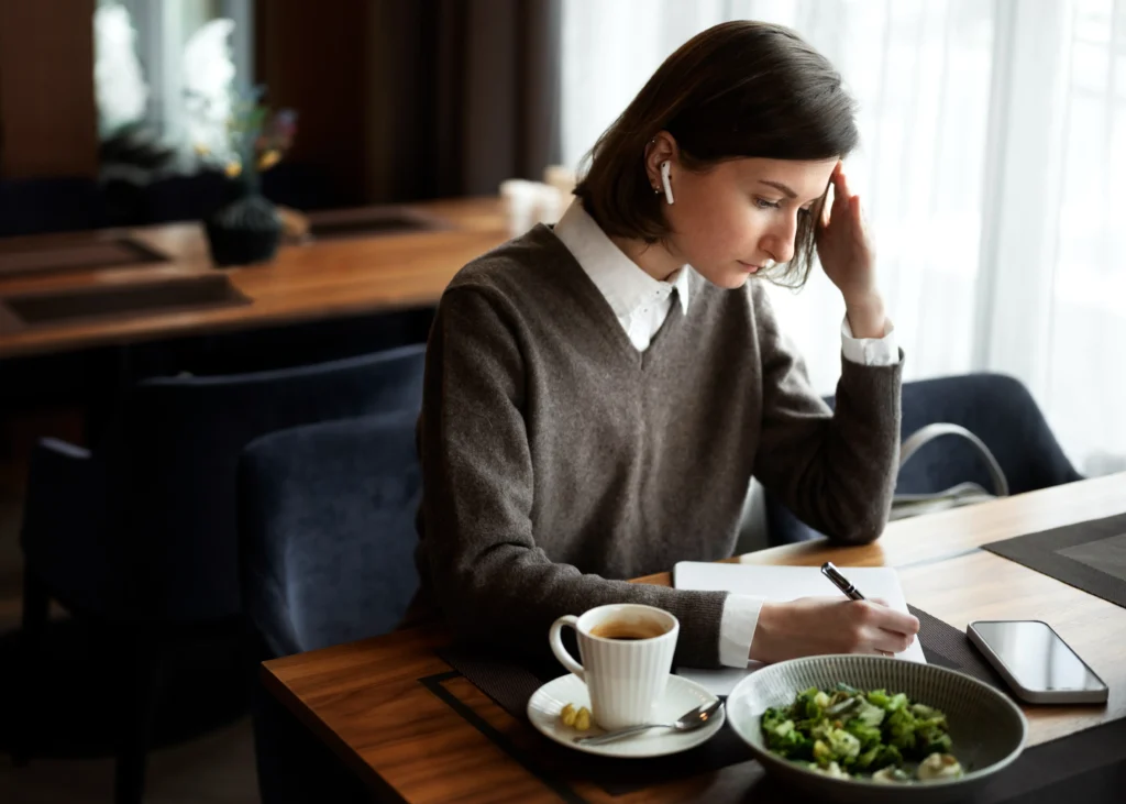 A woman wearing wireless earbuds sits at a café table, resting her head on her hand while writing attentively in a notebook. On the wooden table in front of her sit a cup of coffee, a smartphone, and a bowl with a salad.