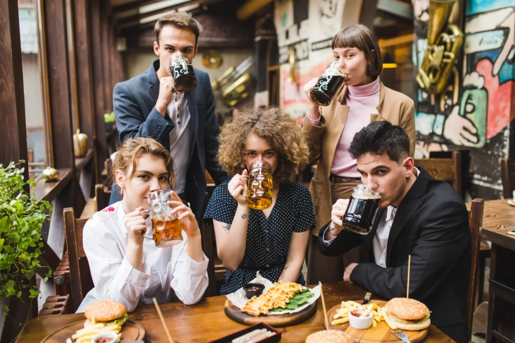Five young adults pose around a wooden restaurant table, each taking a sip simultaneously from large glass steins filled with dark and light beer. Placed on the table in front of them are plates of burgers, French fries, and waffles in an eclectically decorated pub.