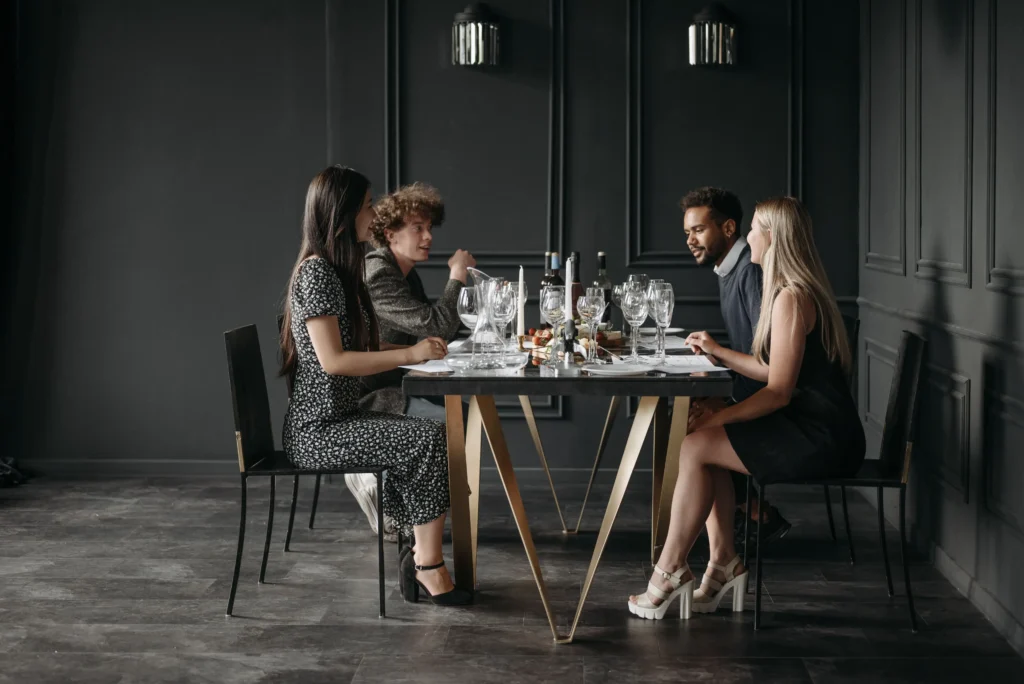 Four people are seated around a modern dining table in a dimly lit, dark gray room with elegant wall molding. They are engaged in conversation during a formal dinner gathering, with wine glasses and table settings arranged in front of them.