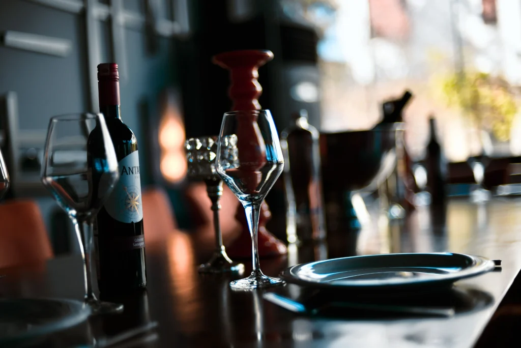 A bottle of Antares red wine sits next to an empty wine glass on a polished wooden table. An elegant plate and additional blurred table settings create a sophisticated restaurant ambiance in the background.