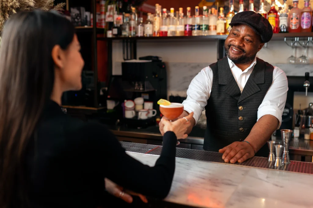 A smiling bartender wearing a flat cap and vest serves a crafted cocktail garnished with a lemon wheel across a marble counter. A female customer seated in the foreground reaches out to accept the drink in a warmly lit bar environment.