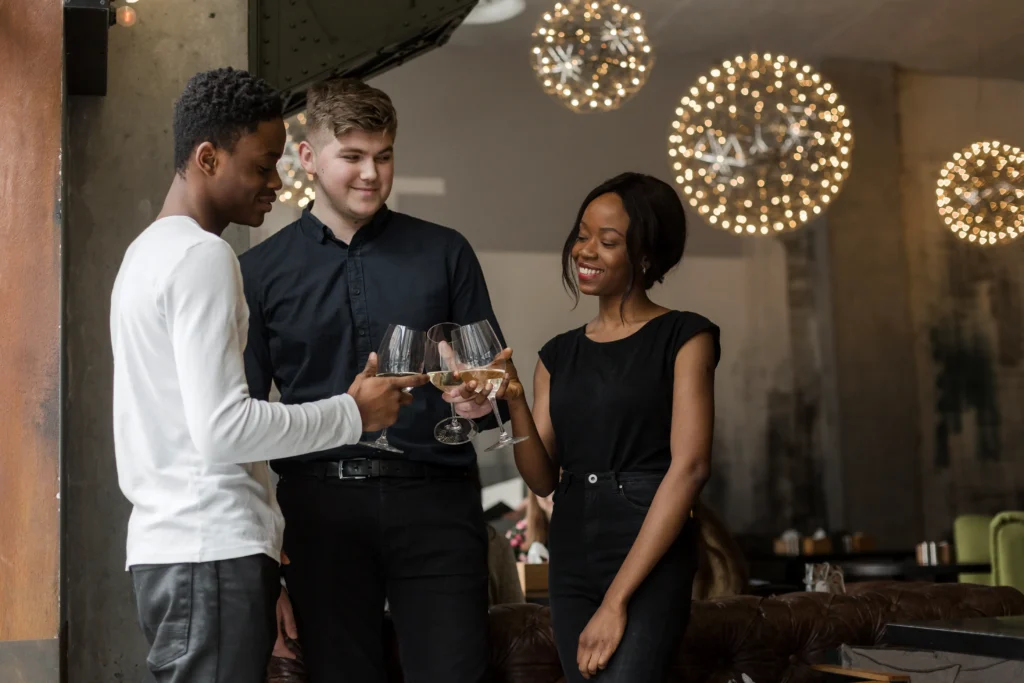 A diverse group of three young friends smile as they gather in a restaurant or bar to clink their wine glasses together in a celebratory toast. In the image, the background features warm, decorative spherical pendant lights glowing softly over an intimate seating area.