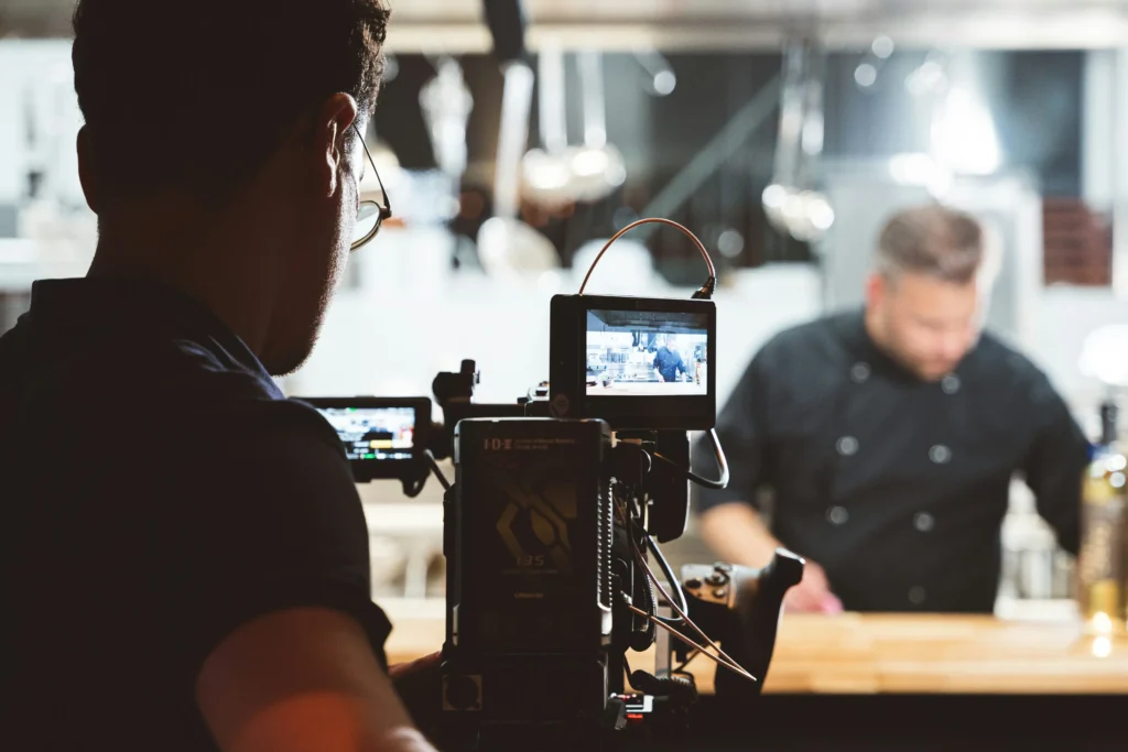 A videographer operates a video camera in a modern kitchen while filming a chef working in the background. The small monitor attached to the camera shows a live preview of the chef preparing food at the counter.