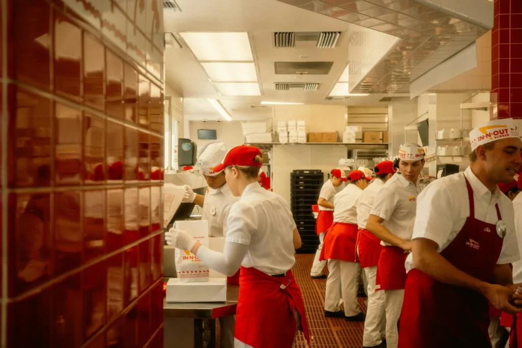 Employees wearing white uniforms, red aprons, and paper hats work bustling inside a brightly lit In-N-Out Burger kitchen. Red tiled walls and overhead lighting surround the staff as they assemble orders and prepare food.