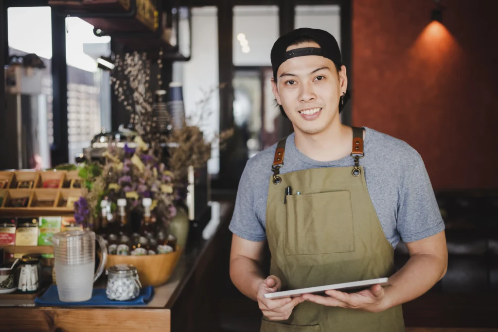 A smiling young male barista wearing a green apron and a backward black cap stands in a coffee shop while holding a digital tablet. The warm cafe background features a wooden counter stocked with jars, dried flowers, and beverage supplies.