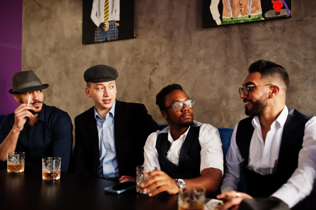A diverse group of four sharply dressed men in retro attire sit together at a table inside a dimly lit venue or club. They are engaged in conversation, holding glasses of whiskey, and wearing classic hats and vests that evoke a vintage mafia or bachelor party aesthetic.