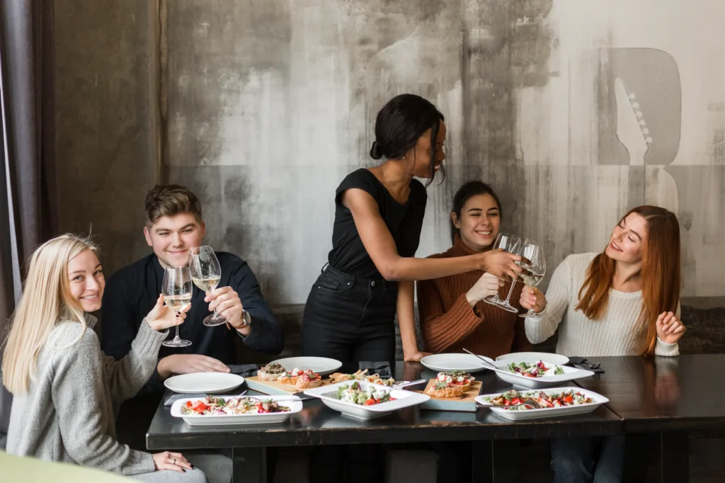 A diverse group of five young adults gathers around a dinner table in a restaurant with a rustic, textured wall background. They are smiling and raising their wine glasses in a celebratory toast over a spread of salads and bruschetta.