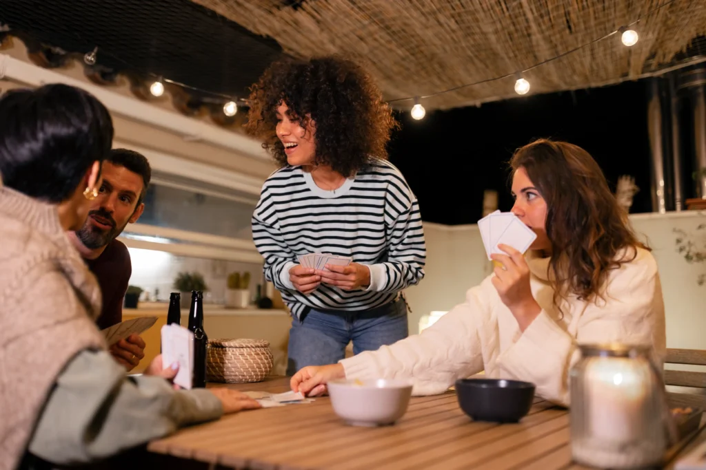 A group of friends gathers around a cozy outdoor table at night, enjoying a card game illuminated by warm string lights. One smiling woman in a striped sweater stands above the table holding her hand of cards while interacting with the others.