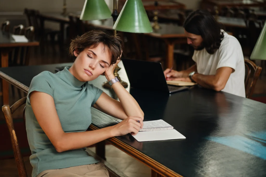 A young woman with short brown hair sleeps while resting her head on her hand at a long wooden table in a library. She holds a pen over an open notebook, while another student works on a laptop in the background.