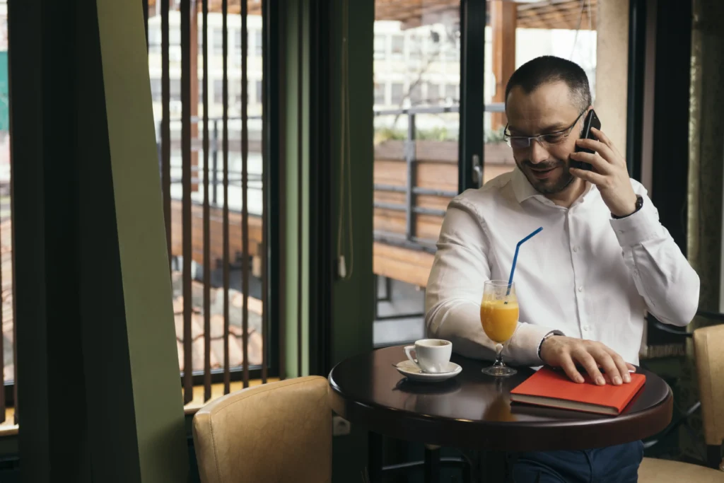 A man wearing a white button-down shirt and glasses sits at a cafe table while talking on his smartphone. On the small table in front of him are an espresso cup, a glass of juice with a blue straw, and a closed red notebook.