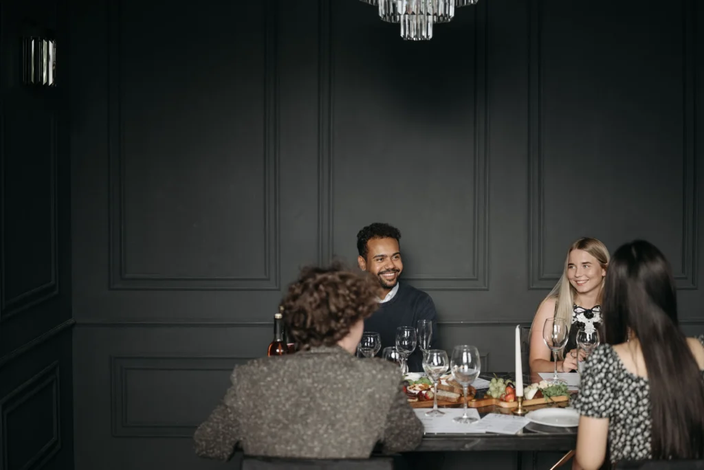 A small group of friends is seated around a dinner table, smiling and conversing during a meal in a room with elegant dark paneled walls. The table is set with wine glasses, a platter of food, and a lit candle, creating an intimate dinner party atmosphere.