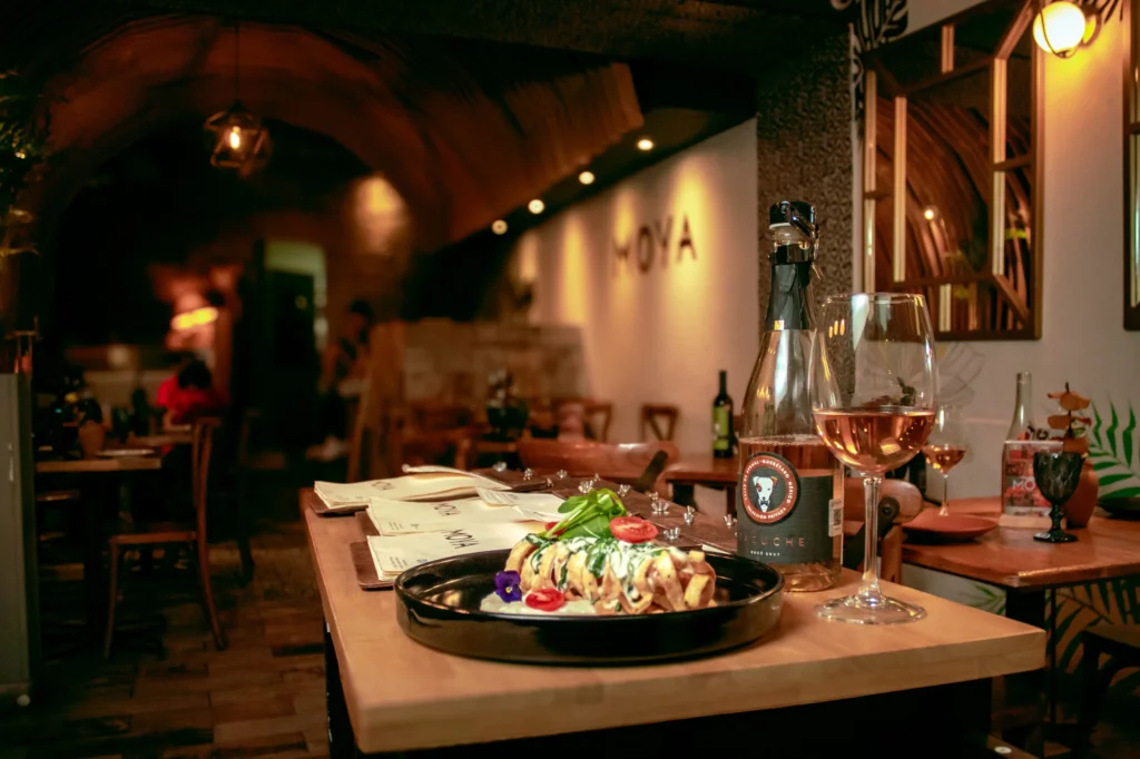 A beautifully plated culinary dish sits on a wooden table alongside a bottle and a glass of rosé wine. The setting is a dimly lit, cozy restaurant featuring warm overhead lighting, wooden furniture, and a background wall with the word "MOYA" on it.