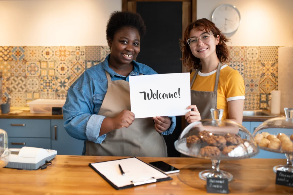 Two smiling women stand behind a cozy bakery counter wearing matching beige aprons while together holding up a white sign that says "Welcome!". Freshly baked pastries in glass display stands and a cash register sit on the wooden counter in front of them.