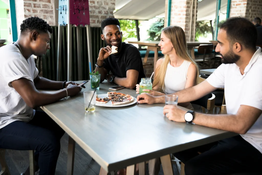 A diverse group of four friends is gathered around a restaurant table with pizza and drinks. While three of them smile and engage in conversation, one man on the left remains distracted by his smartphone.