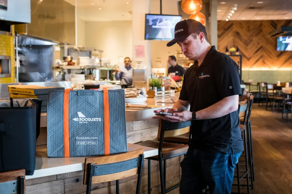 A food delivery driver wearing a FoodJets polo shirt and baseball cap looks at his smartphone inside a restaurant. A large grey branded thermal delivery bag with orange handles sits on the counter beside him.