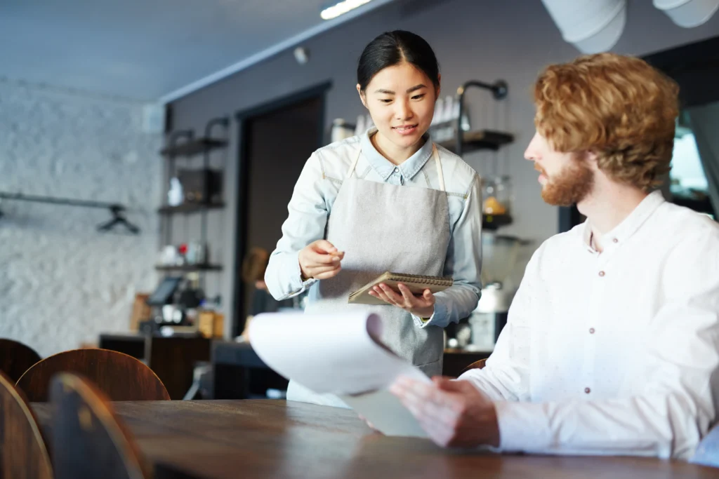 In the image, a waitress wearing a light-blue button-up shirt and a grey apron stands at a wooden table while holding a small notepad. She looks down and smiles gently as she assists a seated male customer with a beard who is holding a menu.