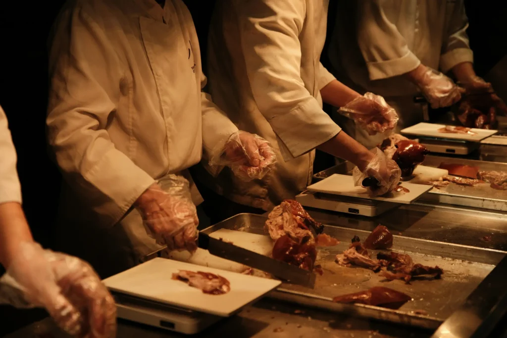 A line of chefs in white jackets and clear plastic gloves carefully carve roasted meat along a metal prep counter. Under warm lighting, each cook places slice portions onto small white plates set atop digital kitchen scales.