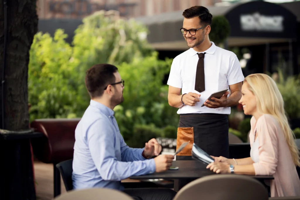 A smiling waiter wearing a white shirt, tie, and apron holds a digital tablet while taking an order from a couple seated at an outdoor restaurant table. The man and woman look up at the waiter warmly while holding menus, with lush green trees blurred in the background.