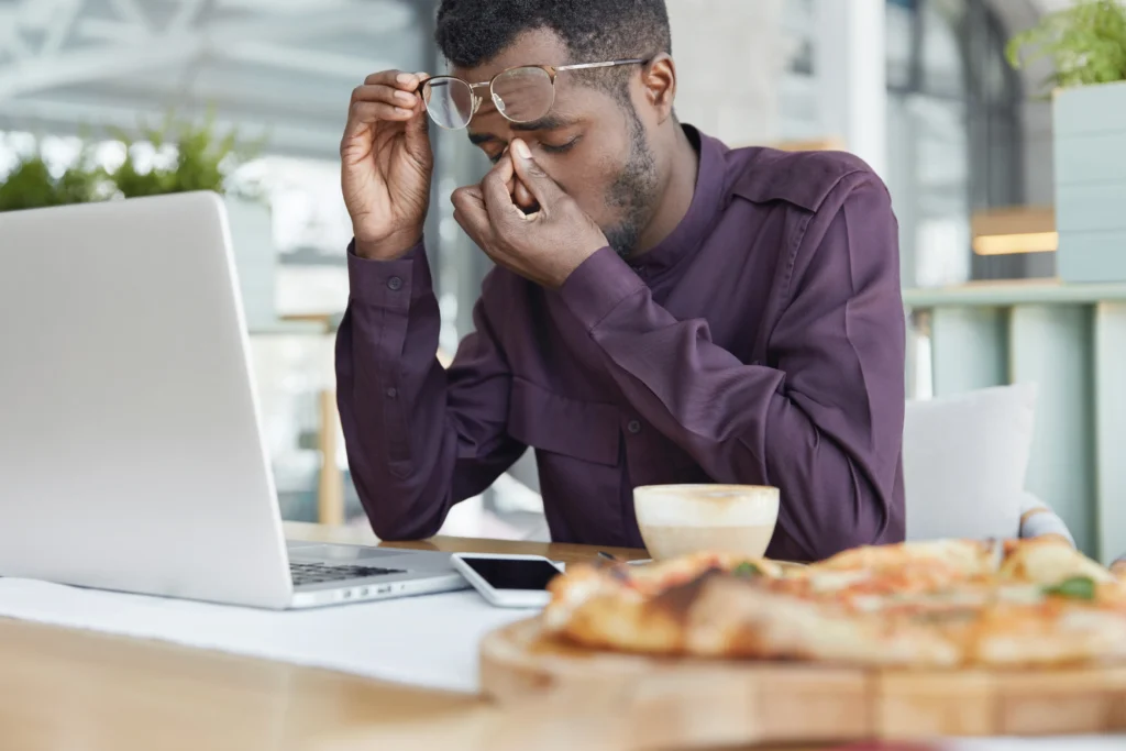 An exhausted man lifts his glasses and pinches the bridge of his nose while sitting in front of an open laptop. His desk is cluttered with a phone, a cup of coffee, and slices of pizza, conveying a sense of fatigue from long hours of work.