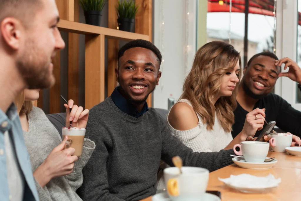 A diverse group of young friends is sitting together at a wooden table in a cozy café, smiling and chatting over coffee and lattes. One woman looks at her smartphone while the others engage in conversation, enjoying their shared hangout.