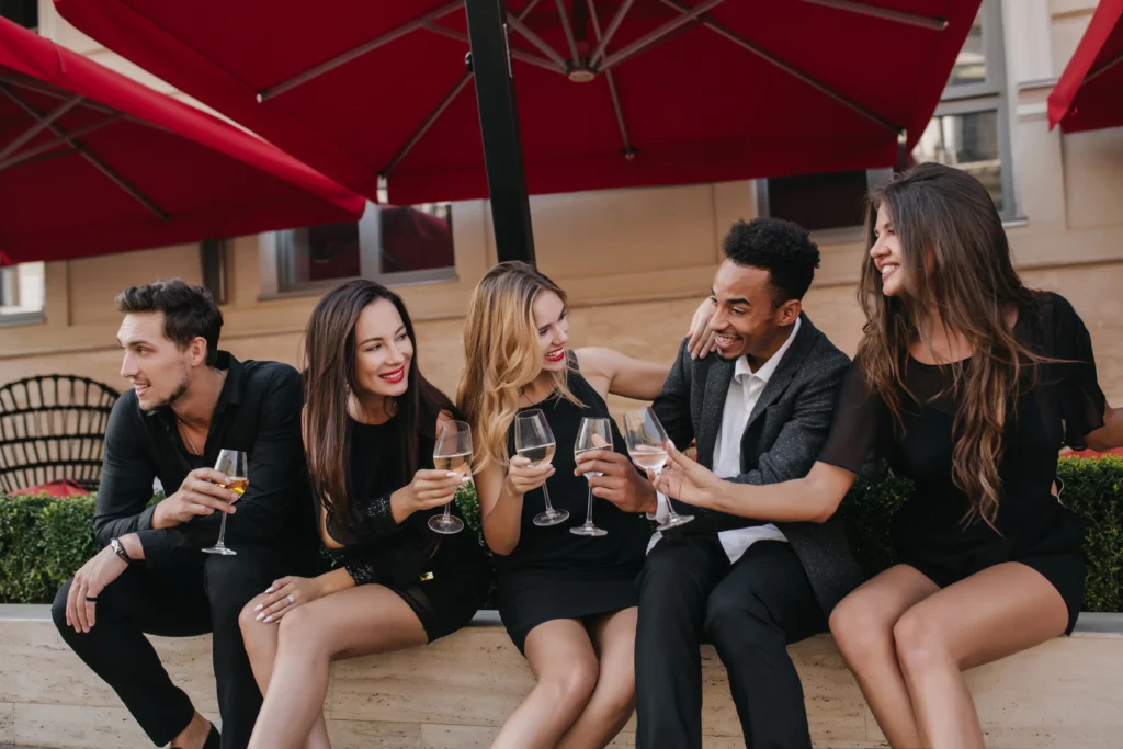 A group of five stylish, smiling friends sit together outside an upscale venue under large red umbrellas while wearing elegant black outfits. They laugh and chat as they raise glasses of white wine or champagne to toast their gathering.
