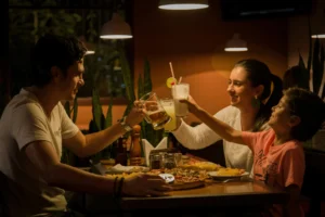 A family sits together at a dimly lit restaurant table filled with pizza, clinking their glasses together in a toast. The father holds a mug of beer, the mother holds a cocktail, and the young child enthusiastically reaches up to join in with his drink.