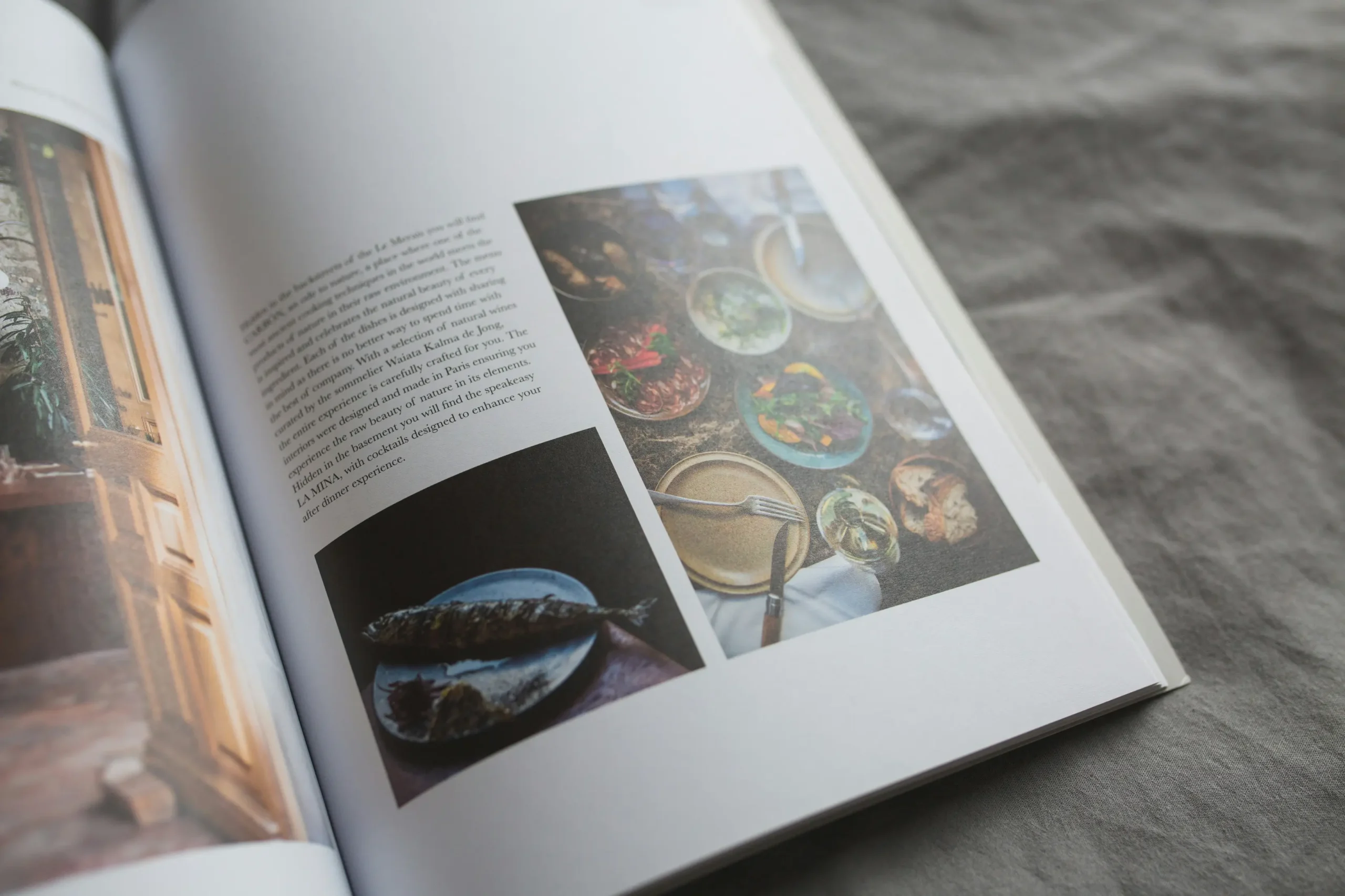 An open magazine lies on a textured gray fabric surface, featuring pages with printed text and food photography. The right page shows a top-down view of a table spread with various small dishes alongside a lower image of a cooked fish on a plate.