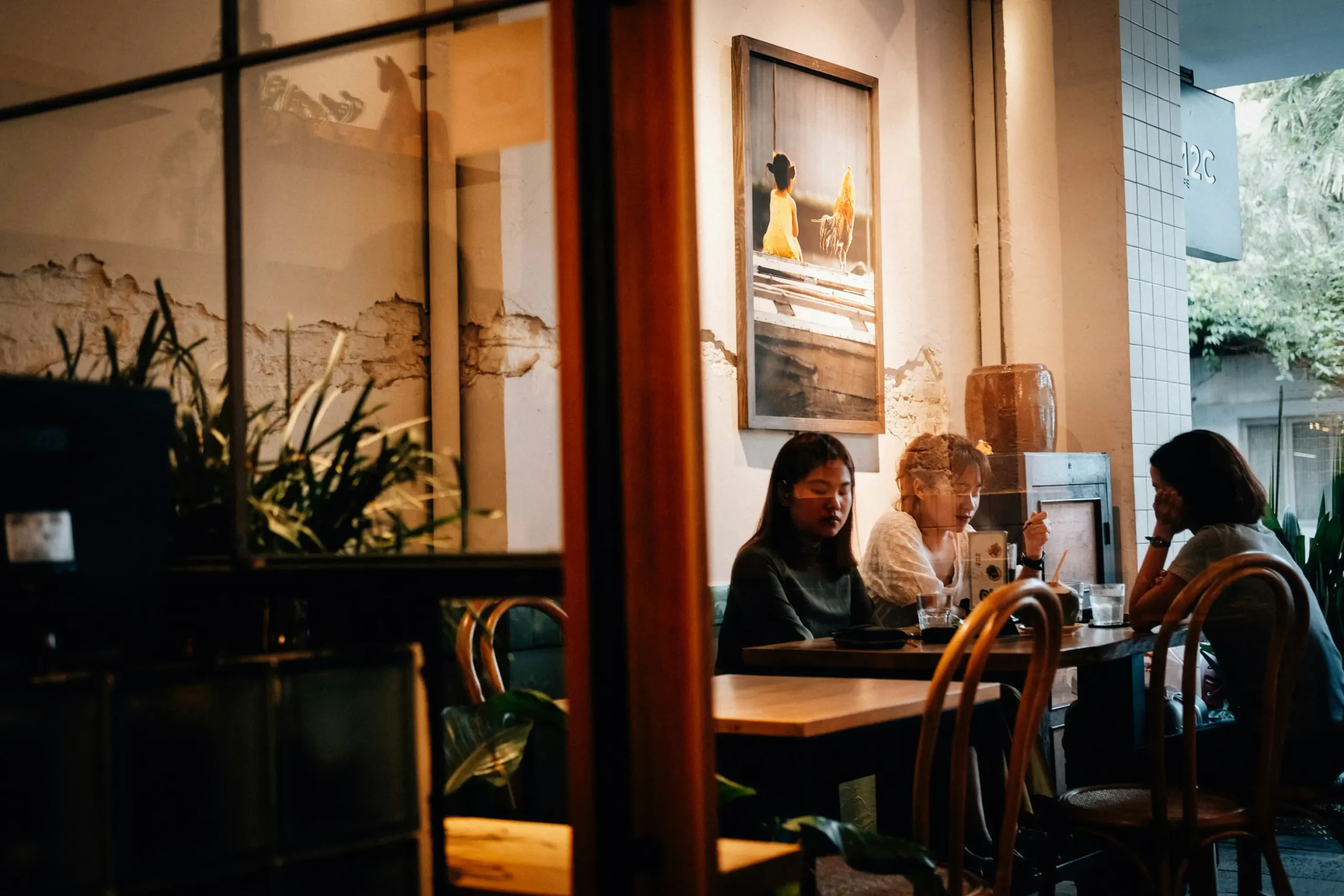Three people sit around a wooden table inside a cozy, dimly lit cafe, seen through a glass window frame. Warm lighting illuminates the rustic interior, featuring a framed artwork on the distressed wall and potted plants nearby.