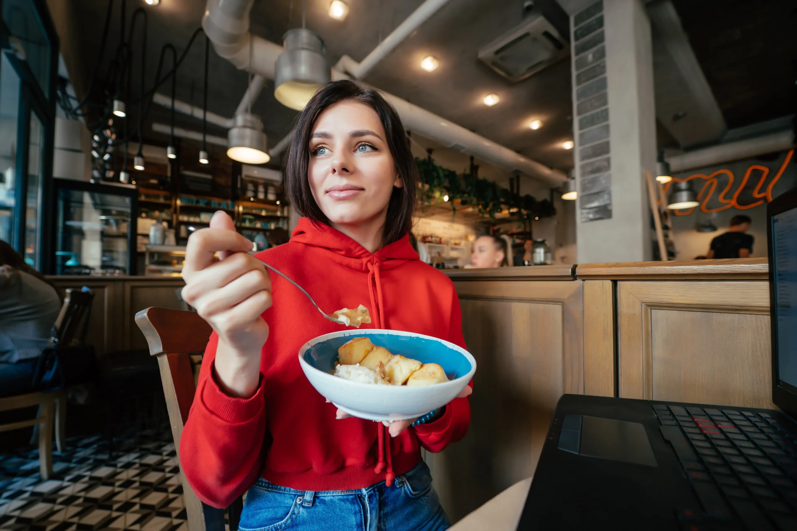A young woman wearing a red hoodie sits at a café table, enjoying a bowl of food with a spoon. A open laptop sits on the table beside her in the brightly lit interior.