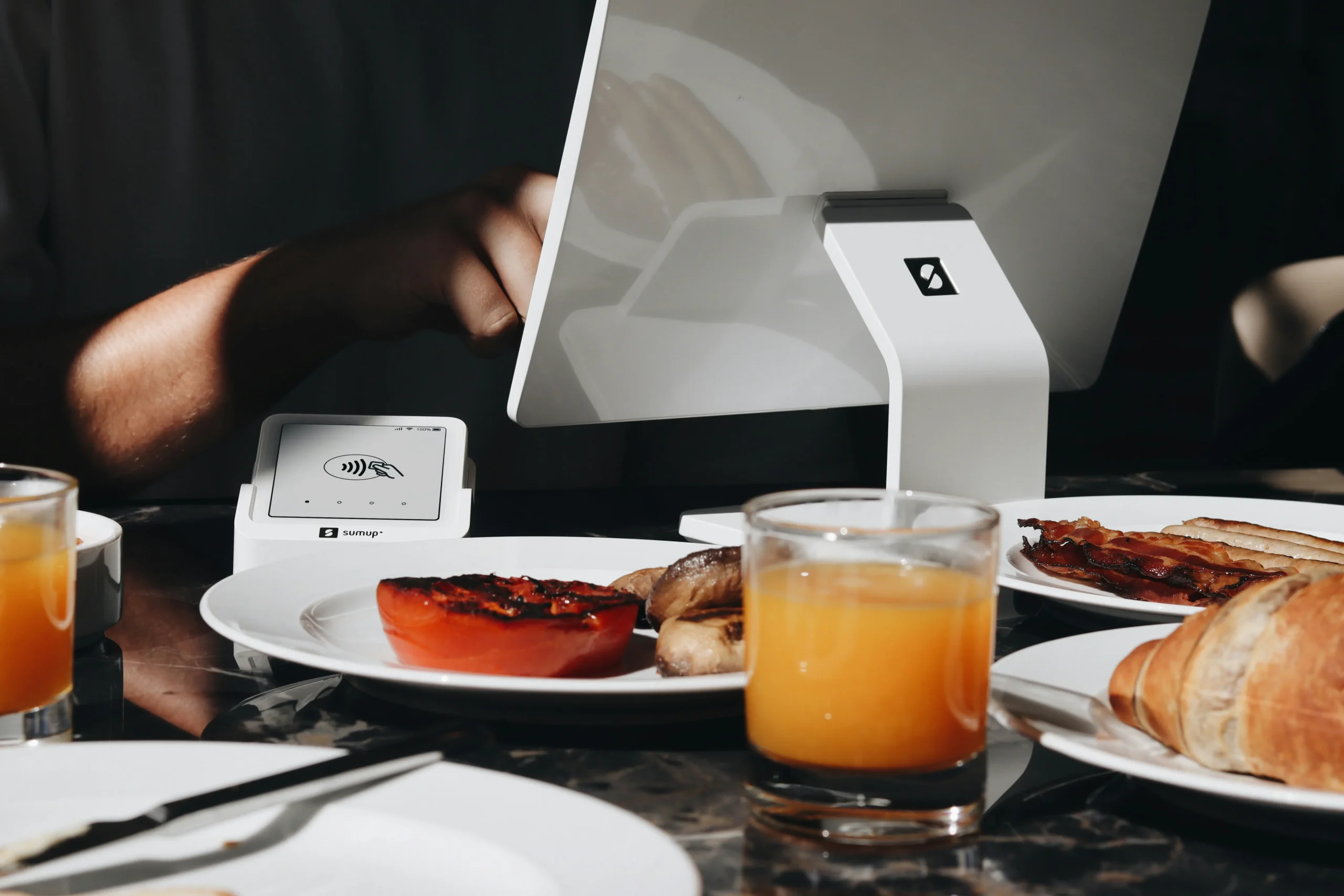 A customer uses a white tablet checkout terminal next to a contactless SumUp card reader at a restaurant table. The table is set with breakfast items, including orange juice, croissants, bacon, and grilled vegetables.