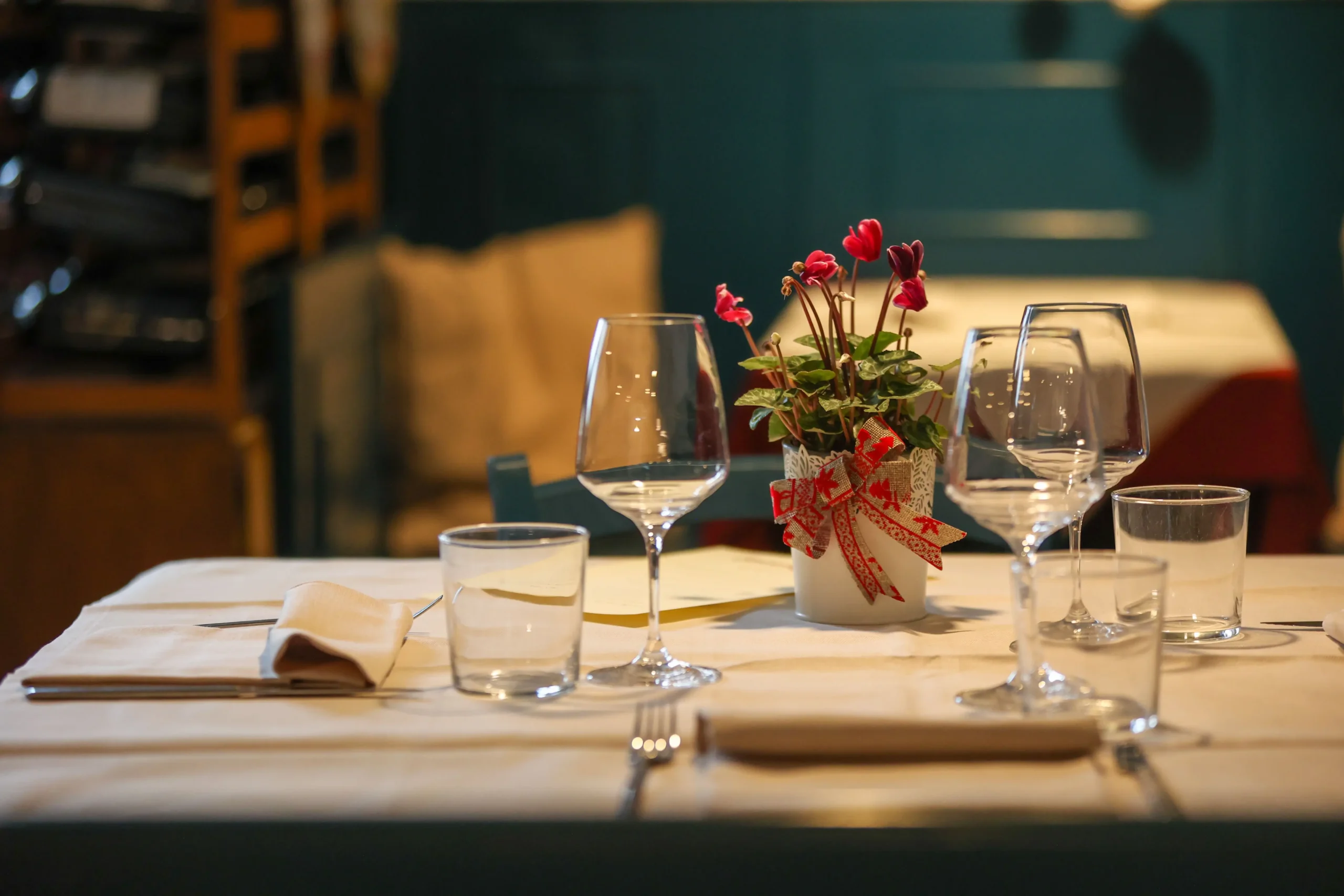 An elegantly set dining table in a cozy restaurant features clear wine glasses, water tumblers, and neatly folded napkins. A small potted plant with vibrant pinkish-red flowers and a decorative red ribbon serves as a charming centerpiece.