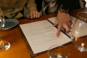 A person points their finger at a item on an open menu placed on a wooden table. Empty wine glasses and small plates sit nearby during a meal.