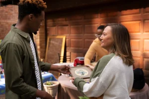 A young man and a woman hold a small framed floral picture together at an outdoor yard sale. The woman smiles as she presents the item to the man outside a wooden garage door.