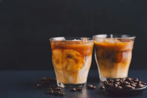 Two small clear glasses filled with iced coffee sit against a dark background, showcasing swirling cream mixing into the dark brew. Roasted coffee beans are scattered across the dark surface around the glasses, with a small dish full of beans in the foreground.