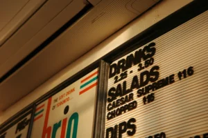 A close-up, angled shot of a retro menu board featuring raised black lettering for drinks, salads, and dips alongside listed prices. Beside it, a portion of a sign displays the Brio beverage logo with the slogan "THE SOFT DRINK OF TORONTO..." under warm indoor lighting.