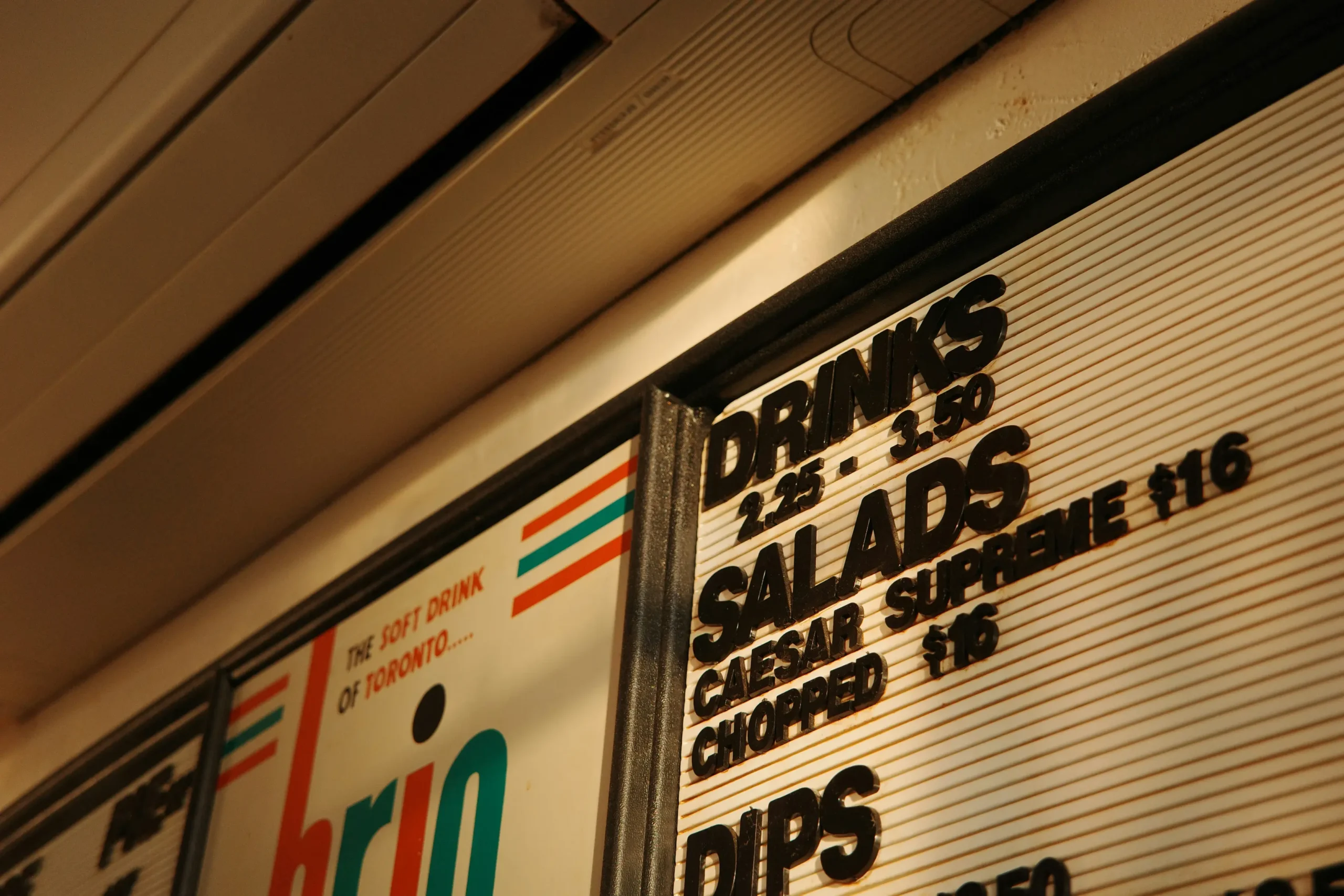 A close-up, angled shot of a retro menu board featuring raised black lettering for drinks, salads, and dips alongside listed prices. Beside it, a portion of a sign displays the Brio beverage logo with the slogan "THE SOFT DRINK OF TORONTO..." under warm indoor lighting.
