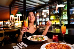 A smiling woman sits at a dimly lit bar table holding a tropical cocktail in one hand and a lit sparkler in the other. In front of her, the table is set with a salad, a pizza, and a second layered cocktail drink.