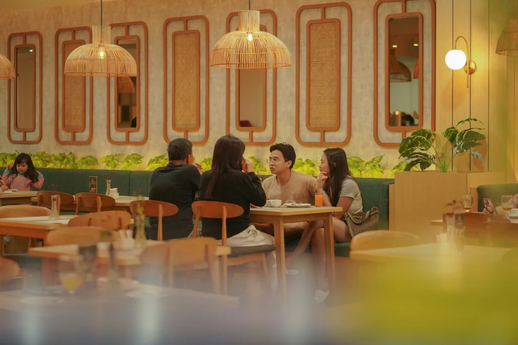 Four people sit around a wooden dining table enjoying a meal together in a warm, modern restaurant. Above them hang woven pendant lights that complement the light wood furniture and decorative wall panels in the background.