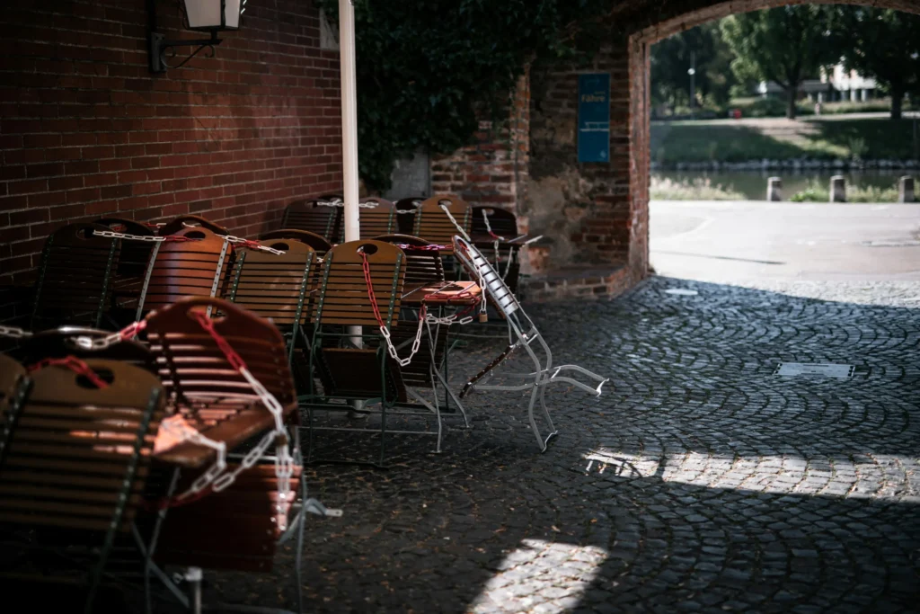 Wooden folding chairs are stacked and secured together with red and white chains next to a brick wall. In the background, a stone archway opens onto a sunlit cobblestone street near a body of water.