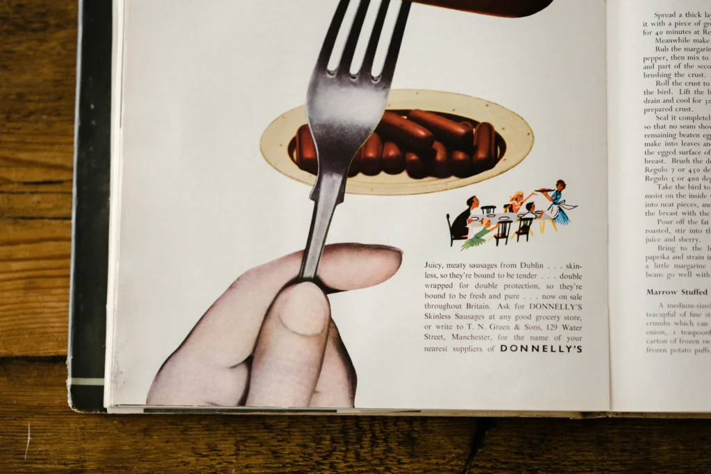 A vintage magazine advertisement features a large hand holding a fork pointed downward over a small plate of sausages. Accompanying the central illustration is descriptive copy for Donnelly's Skinless Sausages alongside a small drawing of a family gathered at a dinner table.