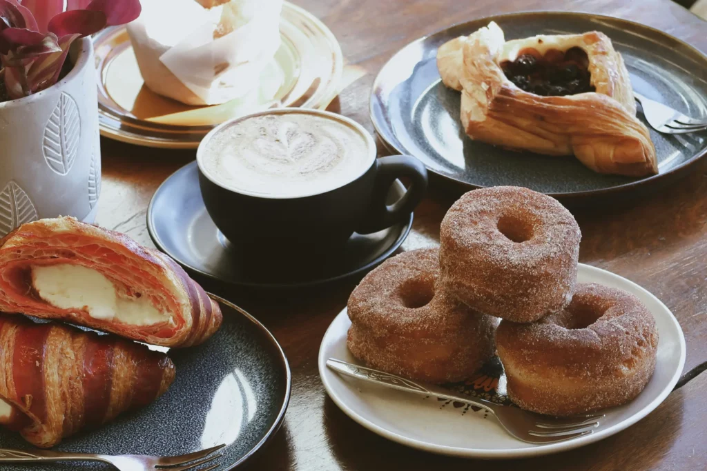 A wooden table is laden with a warm cup of latte art coffee surrounded by an assortment of fresh pastries. The spread features a stack of sugar-dusted donuts, cream-filled striped croissants, a berry Danish, and a potted plant in the background.