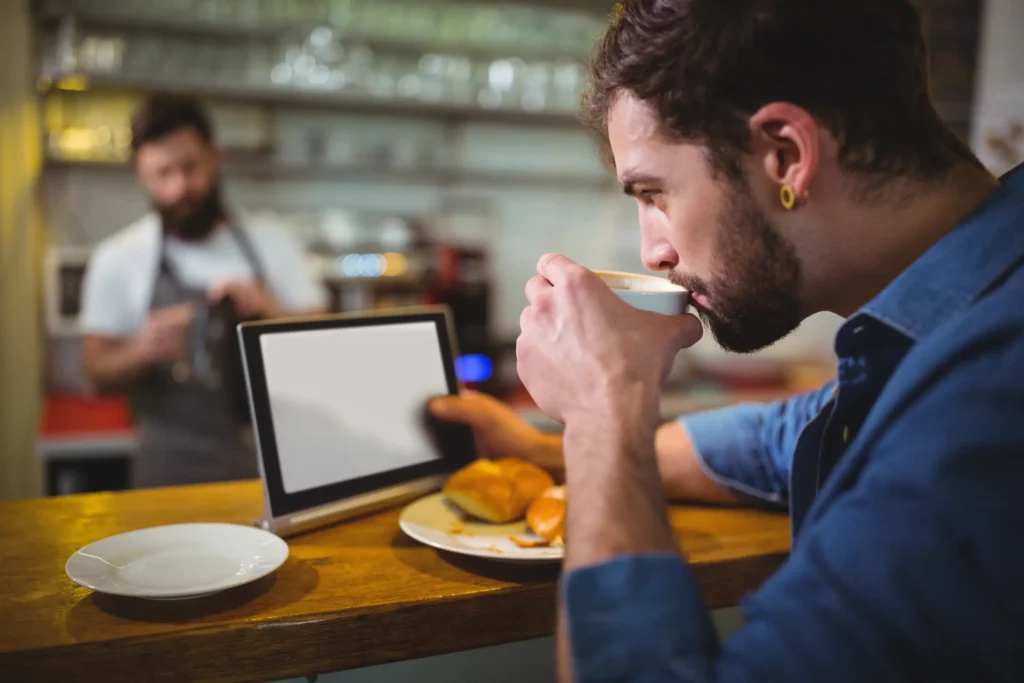 A young bearded man in a blue shirt sits at a wooden coffee shop counter, sipping from a cup while looking at a tablet. On the counter beside his device are a plate of pastries and a small empty dish, with a barista visible in the blurred background.