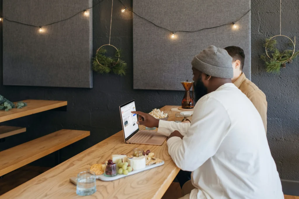 A bearded man in a beanie and white sweater points at a laptop screen while sitting beside a colleague at a long wooden table. Snacks, including a charcuterie board and a bowl of popcorn, are arranged nearby in a dimly lit, cozy cafe environment.
