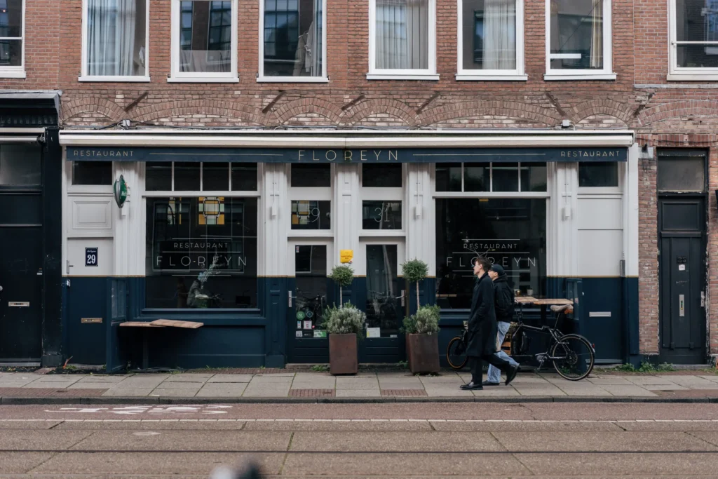 A street-level view shows two pedestrians walking past the dark blue facade of "Restaurant Floreyn" set within a traditional brick building. Small potted plants flank the entrance, and a bicycle is parked beside large window panes along the sidewalk.