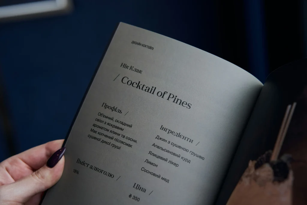 A person holds open a cocktail menu printed on grey paper, showcasing a drink titled "Cocktail of Pines." The page details the flavor profile, alcohol content, price, and ingredients written in Ukrainian.
