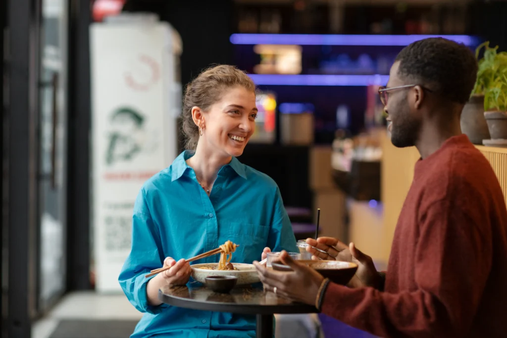 A smiling woman in a bright blue shirt holds noodles with chopsticks as she sits across a small table from a man in a rust-red sweater. They are enjoying a meal together and engaging in a friendly conversation inside a modern restaurant.